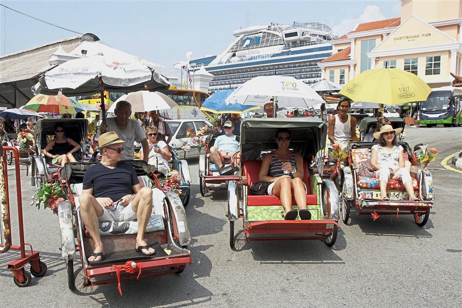 When in Penang: Tourists riding trishaws as a cruise ship towers above in the background in George Town.